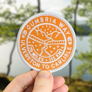 May include: Circular orange patch with white embroidered details. The text reads "CUMBRIA WAY" and "ULVERSTON TO CARLISLE", with "70 MILES 112 KM". The design shows a map of a trail with mountains and a river, ideal for hikers.
