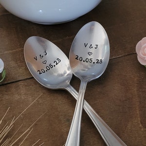 Könnte beinhalten: Zwei silberne Löffel mit gravierten Herzen und dem Text "V & J 20.05.23" auf jedem Löffel. Die Löffel liegen auf einer Holzoberfläche mit einer weißen Teekanne und rosa Blumen im Hintergrund.