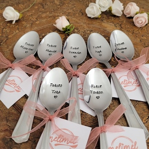 Può includere: Un gruppo di cucchiai d'argento incisi con frasi come "Future Marraine" e "Future Maman", legati con nastri rosa e accompagnati da piccoli biglietti. Rose bianche e altri fiori sullo sfondo.
