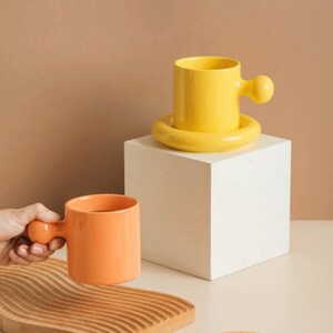 May include: Two ceramic mugs with unique handles, one yellow and one orange, are displayed on a white pedestal and a wooden tray. A small orange saucer with two pieces of bread on it is in the foreground.