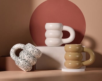 May include: Three ceramic mugs with a unique, rounded, and textured design. The mugs are in white, black and white speckled, and brown.
