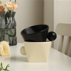 May include: Two ceramic cups, one black and one white, with a rounded handle on each. The cups are stacked on top of each other.