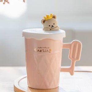 May include: Pink ceramic mug with a bear wearing a crown on the lid. The mug has a handle shaped like an ice cream cone and the text "Make today amazing" on the front.