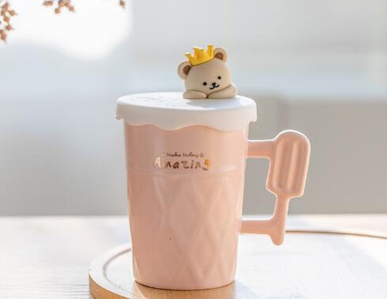 May include: Pink ceramic mug with a bear wearing a crown on the lid. The mug has a handle shaped like an ice cream cone and the text "Make today amazing" on the front.