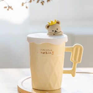 May include: A yellow ceramic mug with a white lid and a small bear figurine on top. The mug has a textured surface and the words "Make today amazing" are printed on the side.