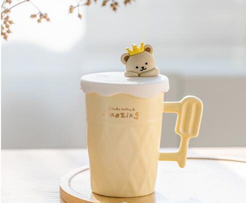 May include: A yellow ceramic mug with a white lid and a small bear figurine on top. The mug has a textured surface and the words "Make today amazing" are printed on the side.
