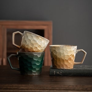 May include: Three ceramic mugs with textured surfaces. The mugs are stacked on top of each other. The top mug is white with brown and beige accents. The middle mug is green. The bottom mug is brown with beige accents. The mugs are sitting on a wooden table with a book.
