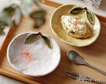 May include: Two small ceramic plates with a peach and a lemon design. The plates have a textured surface and are decorated with green leaves. The lemon plate has a small cookie with white frosting on it. The plates are on a wooden tray with the text "MADE IN JAPAN" on the bottom right corner.