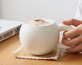 May include: White ceramic mug with a rounded body and a large handle. The mug is filled with a light brown beverage and has a sprinkle of cinnamon on top. The mug is sitting on a brown and white coaster.