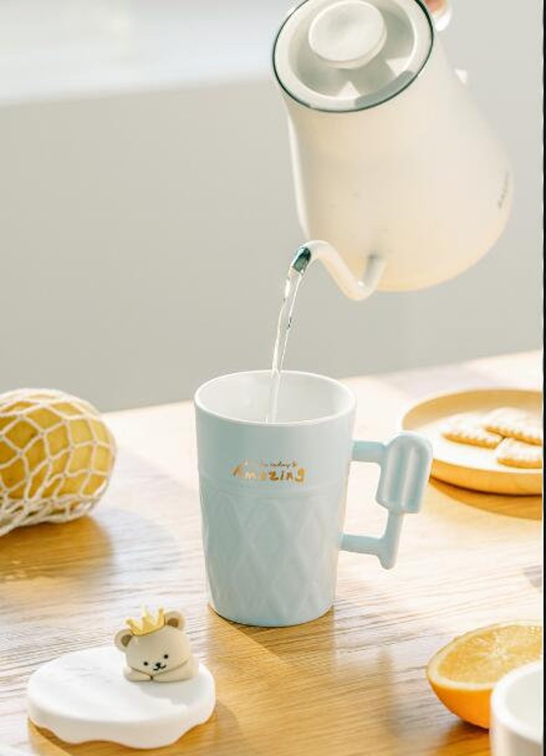 May include: A light blue ceramic mug with a popsicle-shaped handle. The mug has a textured design and the words "everyday amazing" written in gold lettering. The mug is being filled with water from a white teapot.