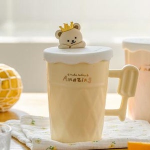 May include: Two ceramic mugs with a bear wearing a crown on the lid. The mugs are yellow and pink and have the text "Make today amazing" on them.
