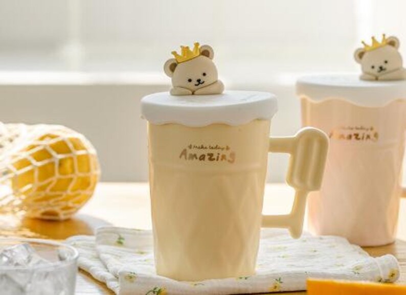 May include: Two ceramic mugs with a bear wearing a crown on the lid. The mugs are yellow and pink and have the text "Make today amazing" on them.