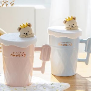 May include: Two ceramic mugs with lids and handles shaped like popsicles. The mugs are decorated with a cartoon bear wearing a crown and the text "Make today amazing". One mug is pink and the other is blue.