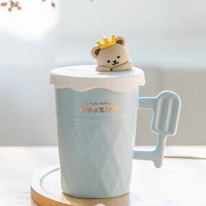 May include: A light blue ceramic mug with a white lid and a bear wearing a crown on top. The mug has a waffle-like texture and a handle shaped like an ice cream popsicle. The mug says "Make today amazing" in gold lettering.