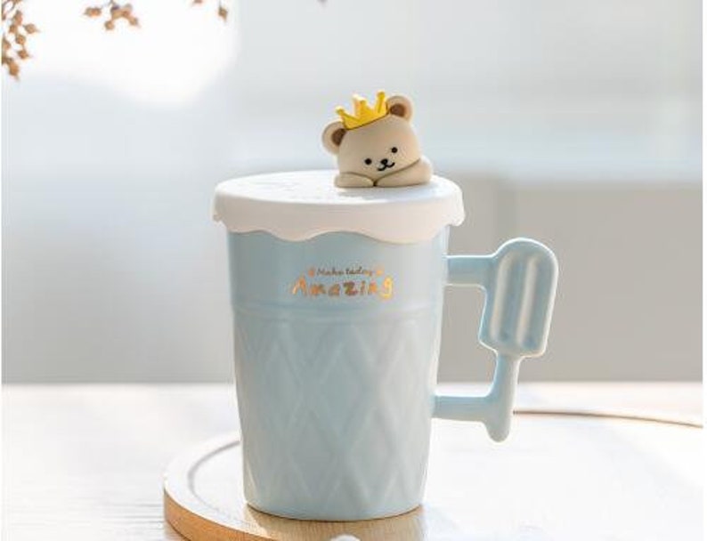 May include: A light blue ceramic mug with a white lid and a bear wearing a crown on top. The mug has a waffle-like texture and a handle shaped like an ice cream popsicle. The mug says "Make today amazing" in gold lettering.