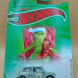 May include: A Hot Wheels toy car featuring a green Grinch theme. The packaging includes the Grinch and his dog, Max, in a Christmas setting. The car is a Two Sizes Too Small Coupe.