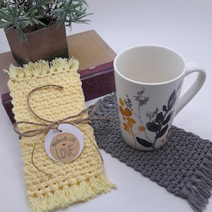 May include: Two crocheted coasters, one yellow with fringe and a tag that says "handmade with love", and one gray with fringe. A white mug with a floral design sits on the gray coaster.