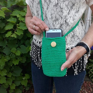 Model wearing an emerald green cell phone purse in a crossbody style. With right hand she is holding a cell phone half way out of the bag to show one way the bag is useful. With the other hand she is holding the bottom of the bag. An outside picture.