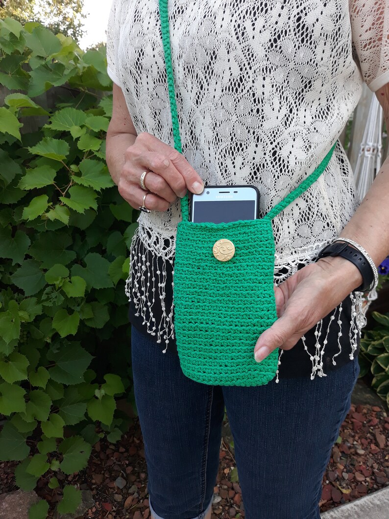 Model wearing an emerald green cell phone purse in a crossbody style. With right hand she is holding a cell phone half way out of the bag to show one way the bag is useful. With the other hand she is holding the bottom of the bag. An outside picture.