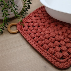 May include: A terracotta crocheted pot holder with a wooden ring handle. The pot holder is on a wooden surface with a white ceramic bowl on top.