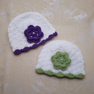 Two white baby hats one under the other placed at an angle. Violet trim and flower and other sage green trim and flower. In middle of the flower a white plastic pearl. Hat stitch has a dainty shell look, soft for a babies little head. So warm!