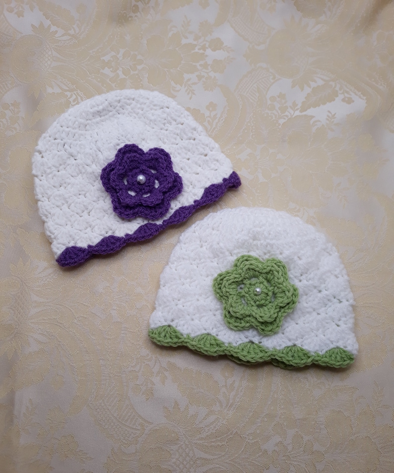 Two white baby hats one under the other placed at an angle. Violet trim and flower and other sage green trim and flower. In middle of the flower a white plastic pearl. Hat stitch has a dainty shell look, soft for a babies little head. So warm!