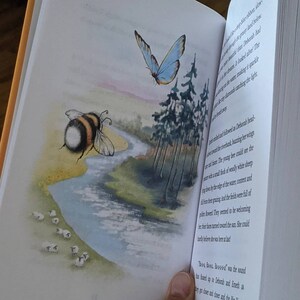 May include: A watercolor illustration of a bee flying over a stream with a flock of white sheep grazing on the bank. A blue butterfly flies above a group of trees in the background. The text reads "The young bee could see the sheep with a small flock of woolly white sheep grazing by the edge of the water, content and full of their flowers. They seemed to be welcoming her, their faces turned toward the sun. She could hardly believe she was here at last!"