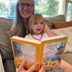 May include: A woman and a young girl are sitting on a couch reading a book titled "Deborah and Kite at the Big River". The book is open and the pages are yellow. The woman is wearing glasses and has gray hair. The girl is wearing a pink shirt and a pink necklace.