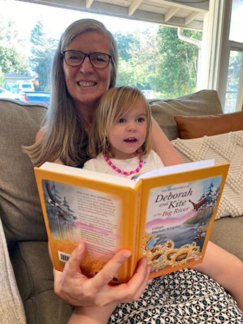 May include: A woman and a young girl are sitting on a couch reading a book titled "Deborah and Kite at the Big River". The book is open and the pages are yellow. The woman is wearing glasses and has gray hair. The girl is wearing a pink shirt and a pink necklace.