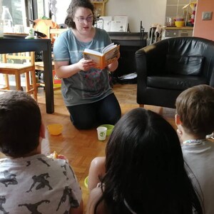 May include: A woman reads a book to three children sitting on the floor. The woman is wearing a grey shirt and jeans. The children are listening attentively. There are two yellow bowls and a green cup on the floor.