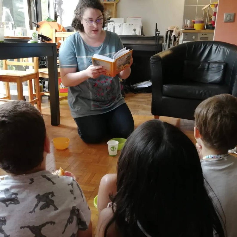May include: A woman reads a book to three children sitting on the floor. The woman is wearing a grey shirt and jeans. The children are listening attentively. There are two yellow bowls and a green cup on the floor.