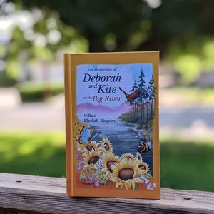 May include: A yellow book titled "The Adventures of Deborah and Kite at the Big River" by Gillian Blackah-Kingsley. The book cover features an illustration of a river, sunflowers, a bee, and a blue butterfly.