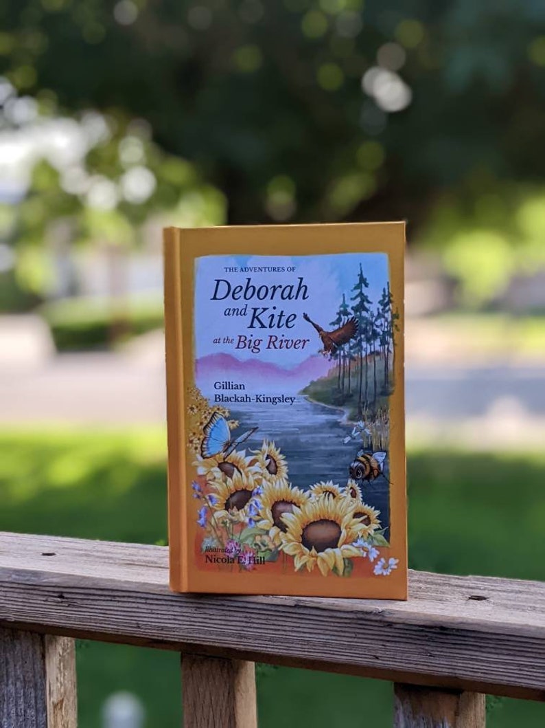 May include: A yellow book titled "The Adventures of Deborah and Kite at the Big River" by Gillian Blackah-Kingsley. The book cover features an illustration of a river, sunflowers, a bee, and a blue butterfly.