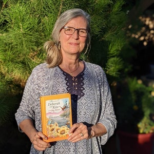 May include: A woman is holding a book titled "Deborah and Kite at the Big River" by Deborah Kingsley. The book cover features a colorful illustration of a river, a kite, and sunflowers.