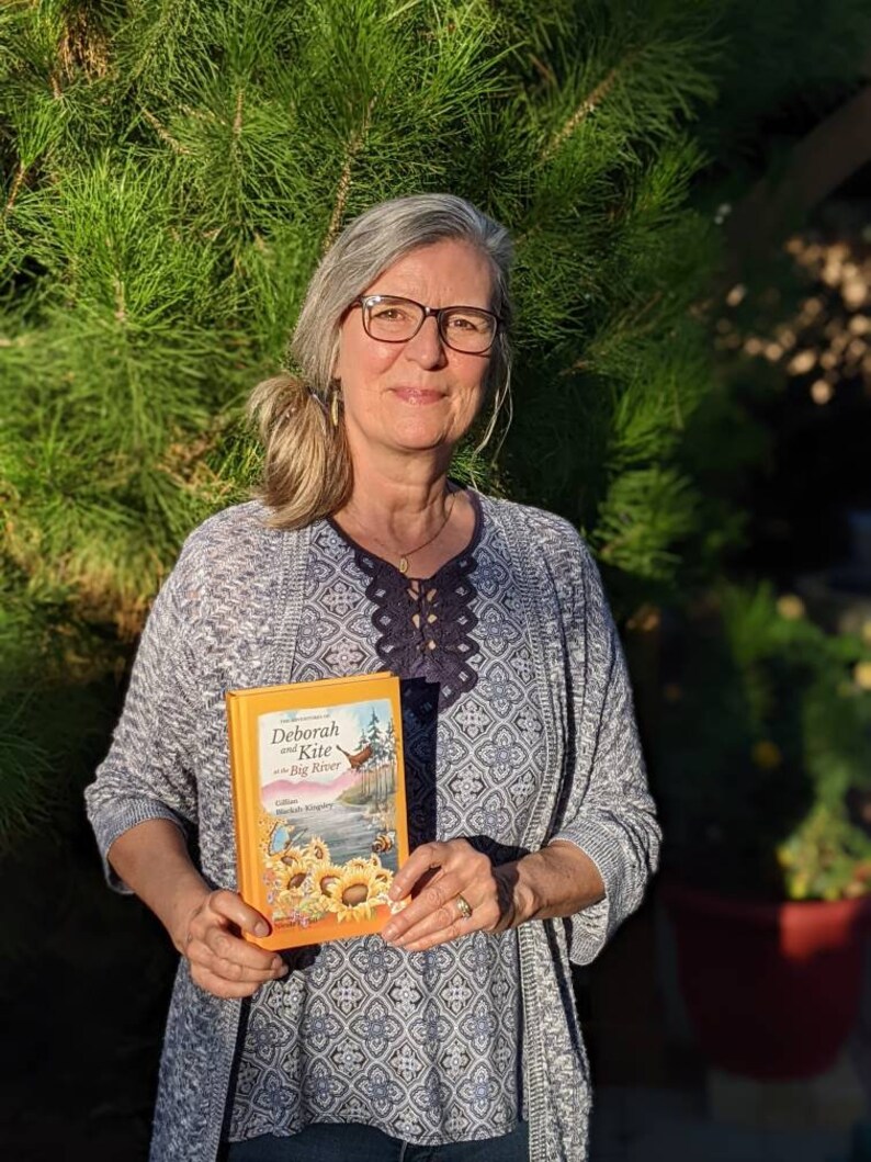 May include: A woman is holding a book titled "Deborah and Kite at the Big River" by Deborah Kingsley. The book cover features a colorful illustration of a river, a kite, and sunflowers.