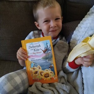 May include: A young child is sitting on a couch, holding a book titled "The Adventures of Deborah and Kite at the Big River" by Gillian Blackah-Kingsley. The child is also holding a peeled banana.