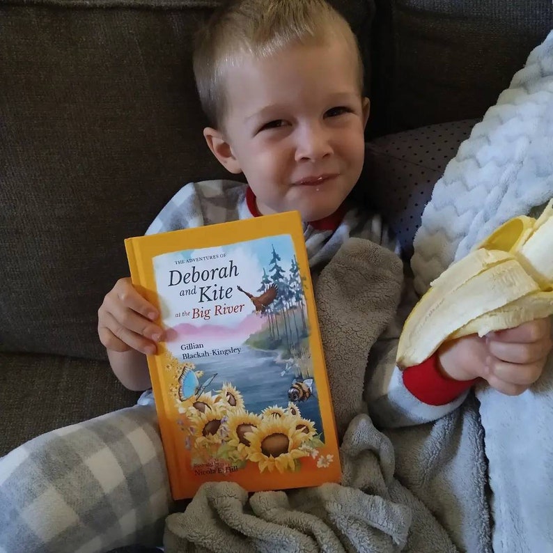 May include: A young child is sitting on a couch, holding a book titled "The Adventures of Deborah and Kite at the Big River" by Gillian Blackah-Kingsley. The child is also holding a peeled banana.