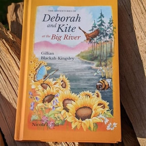 May include: An orange book cover with the title "The Adventures of Deborah and Kite at the Big River" by Gillian Blackah-Kingsley. The cover features illustrations of a river, sunflowers, a bee, a dragonfly, and a bird flying over a forest.
