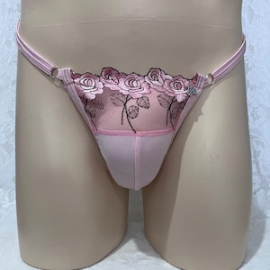 Op de afbeelding: Een lichtroze string met een transparant mesh paneel aan de voorzijde, versierd met geborduurde roze rozen en zwarte stelen. Het kledingstuk heeft dunne elastische zijbandjes die verbonden zijn door zilverkleurige metalen ringen en een kleine hartvormige bedel.