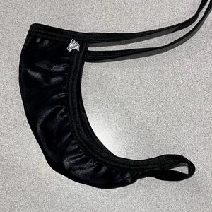 Può includere: Intimo nero satinato con un'emblema argentata. L'intimo presenta un design curvo con spalline sottili e fasce elastiche. Il tessuto ha una leggera lucentezza e lo stile generale è minimalista.