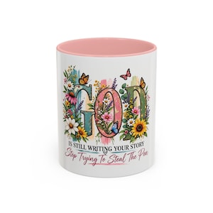May include: A white ceramic mug with a soft pink interior. The exterior features the word GOD in large, floral-filled letters, surrounded by butterflies and wildflowers. Below, the text reads: IS STILL WRITING YOUR STORY Stop Trying To Steal The Pen.