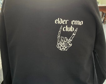 Puede incluir: Sudadera negra de manga larga con un gráfico blanco de una mano de esqueleto haciendo un signo de rock and roll. El texto "elder emo club" está impreso sobre la mano.