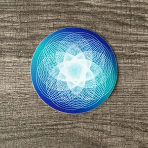 May include: A round sticker with a blue to teal gradient background. A white geometric design with a starburst pattern is centered on the sticker.