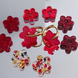 May include: A selection of flower-shaped earrings and charms in shades of red and clear with gold hoops. Some flowers are solid red, others are glittery, and some are clear with colourful accents.