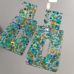 May include: Clear rectangular earrings with a rectangular top, filled with teal, blue, and gold glitter. The earrings have a cutout rectangle in the center of the larger rectangle. The earrings hang from gold-colored hooks.