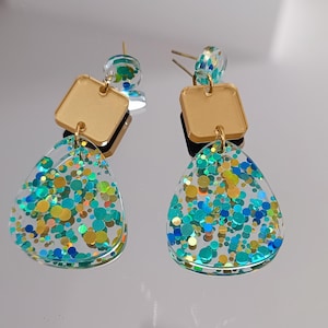 Peut inclure: Une paire de boucles d'oreilles en résine transparente et dorée avec une forme de larme et des accents carrés dorés. Les formes de larme sont remplies de paillettes bleues, vertes et dorées irisées.