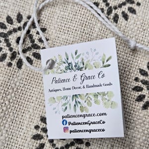 May include: A white tag with the text "Patience & Grace Co" and floral illustrations. The tag is attached to a white string. The tag also includes the website and social media handles.