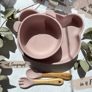 Könnte beinhalten: Rosa-farbenes Silikon-Teller- und Schüssel-Set in Bärenform mit Saugnapf. Das Set enthält zwei Holzlöffel, wobei einer den Namen "Mathilda" eingraviert hat. Der Text "BPA-frei ohne Weichmacher" und "mit Saugnapf" ist auf dem Bild zu sehen. Der Text "in 4 Farben" ist ebenfalls sichtbar.