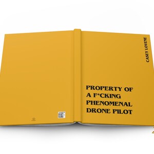 May include: A yellow hardcover book with black text on the front cover that reads PROPERTY OF A F*CKING PHENOMENAL DRONE PILOT and CASEY LOVENE on the spine area. The back cover features a small square barcode and a tiny logo at the bottom.