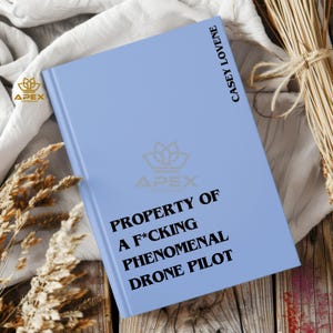 May include: A light blue hardcover book with black text that reads PROPERTY OF A F*CKING PHENOMENAL DRONE PILOT on the front cover and CASEY LOVENE on the spine area. The book features a faint logo in the center and a small logo in the corner.